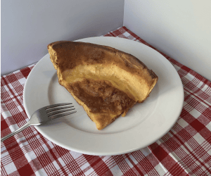 Dutch baby slice on plate with fork, on table with red and white checked tablecloth