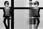 This is a black and white manga illustration showcasing an early design of Sai Ishikawa. The artwork features a mirrored diptych of the character leaning against a black cross, inscribed with vertical katakana characters spelling out his name. A digital glitch effect is applied across the mirrored right side.
