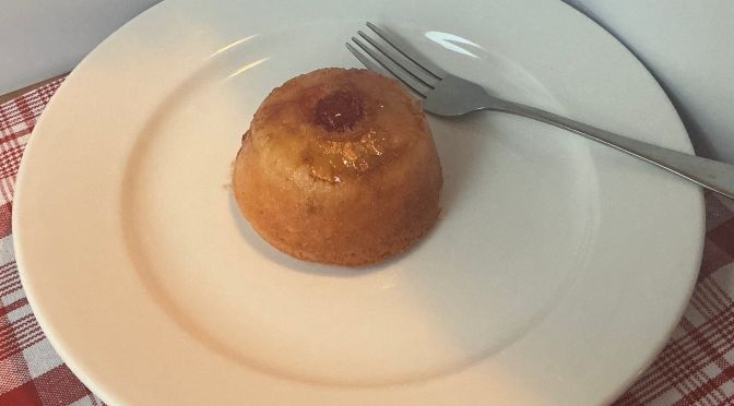 pineapple upside down cake with cherry in center on white plate with a silver fork on it.
