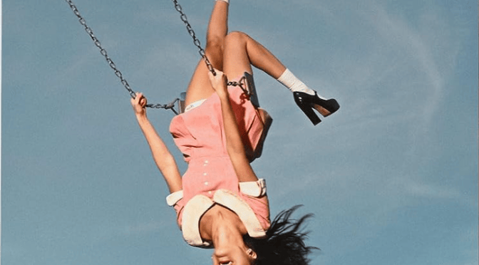 cover of Olivia Rodrigo's album you seem pretty sad for a girl so in love'. shows a girl in a red and white dress on a swing upside down against a blue sky background